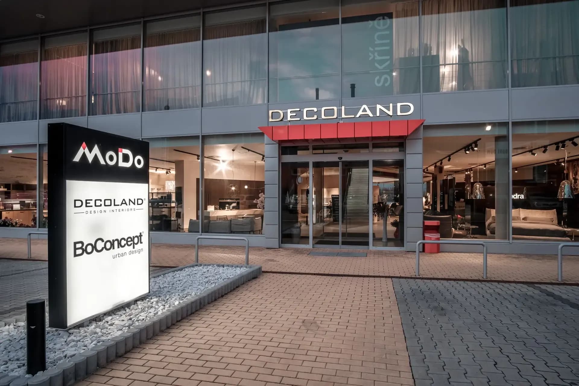 Fotografie fasády showroomu decoland v Průhonicích z pohledu chodníku, v levo reklamní poutač pro decoland MoDO, BoConcept
